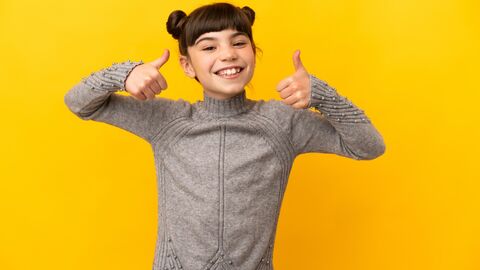 Zwei Daumen hoch! Mädchen mit lustigen Zöpfen steht vor einer gelben Wand. Sie lacht breit und zeigt beide Daumen hoch