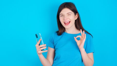 Das ist spitze! Mädchen in blauem Shirt steht vor einer blauen Wand. Sie zwinkert, hält ein Handy in der einen Hand und macht ein TOP Zeichen mit der anderen