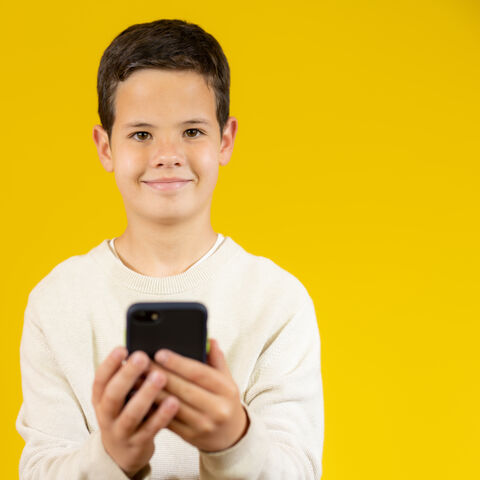 Wissen und Spaß auf dem Smartphone Junge vor gelben Hintergrund hält ein Handy in beiden Händen. Er lächelt in die Kamera.